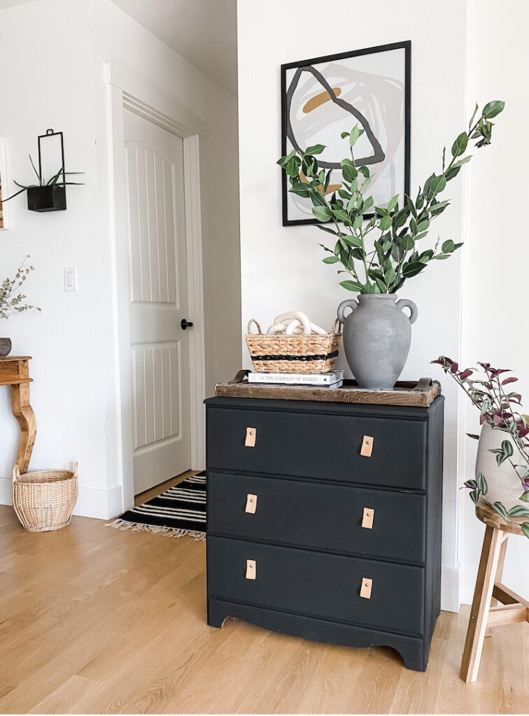 styled home space with painted black dresser, artwork, baskets, and plant stems in vase